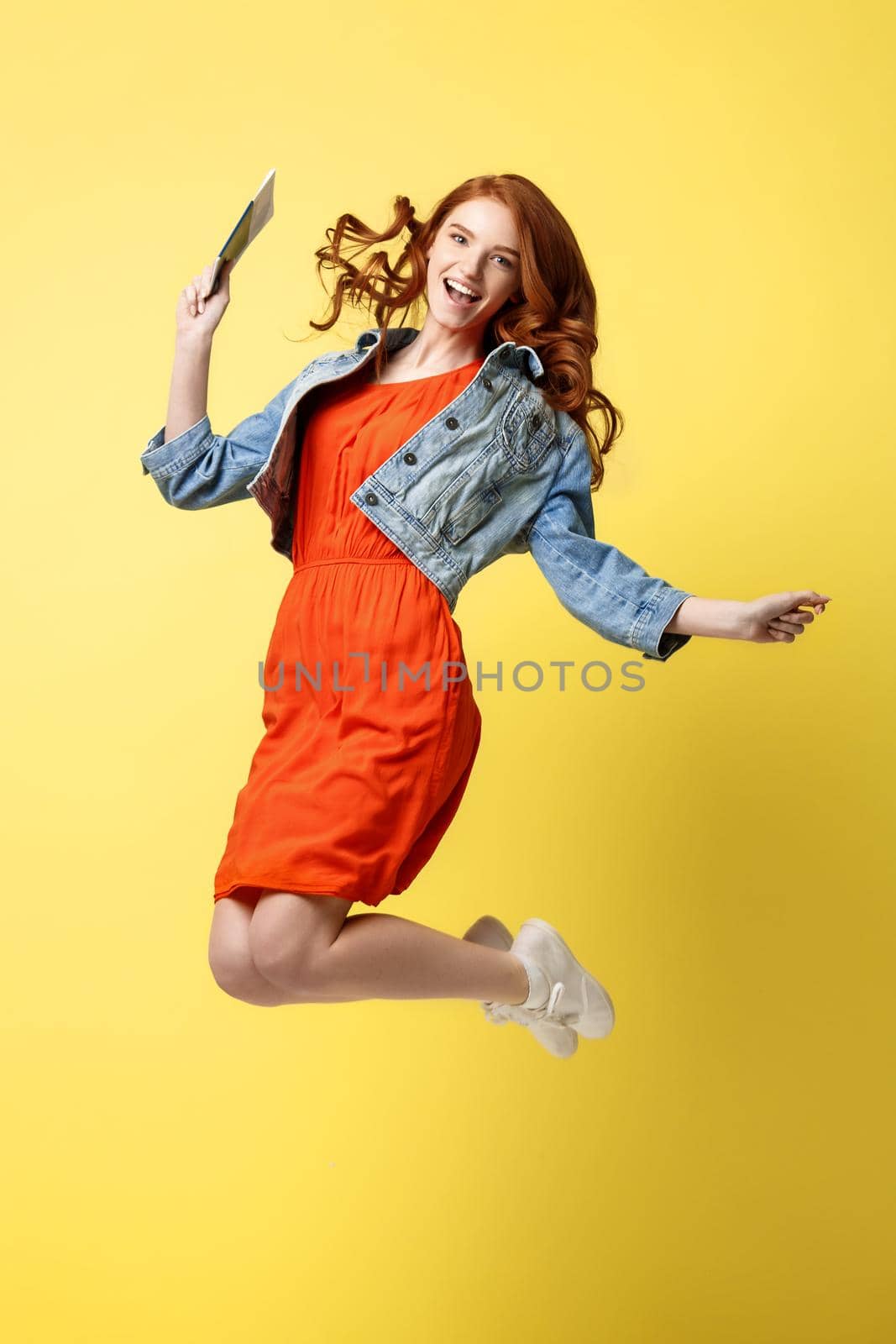 Travel and Lifestyle Concept: Full length portrait red hair happy young woman holding traveling tickets with passport and jumping isolated over bright yellow background.