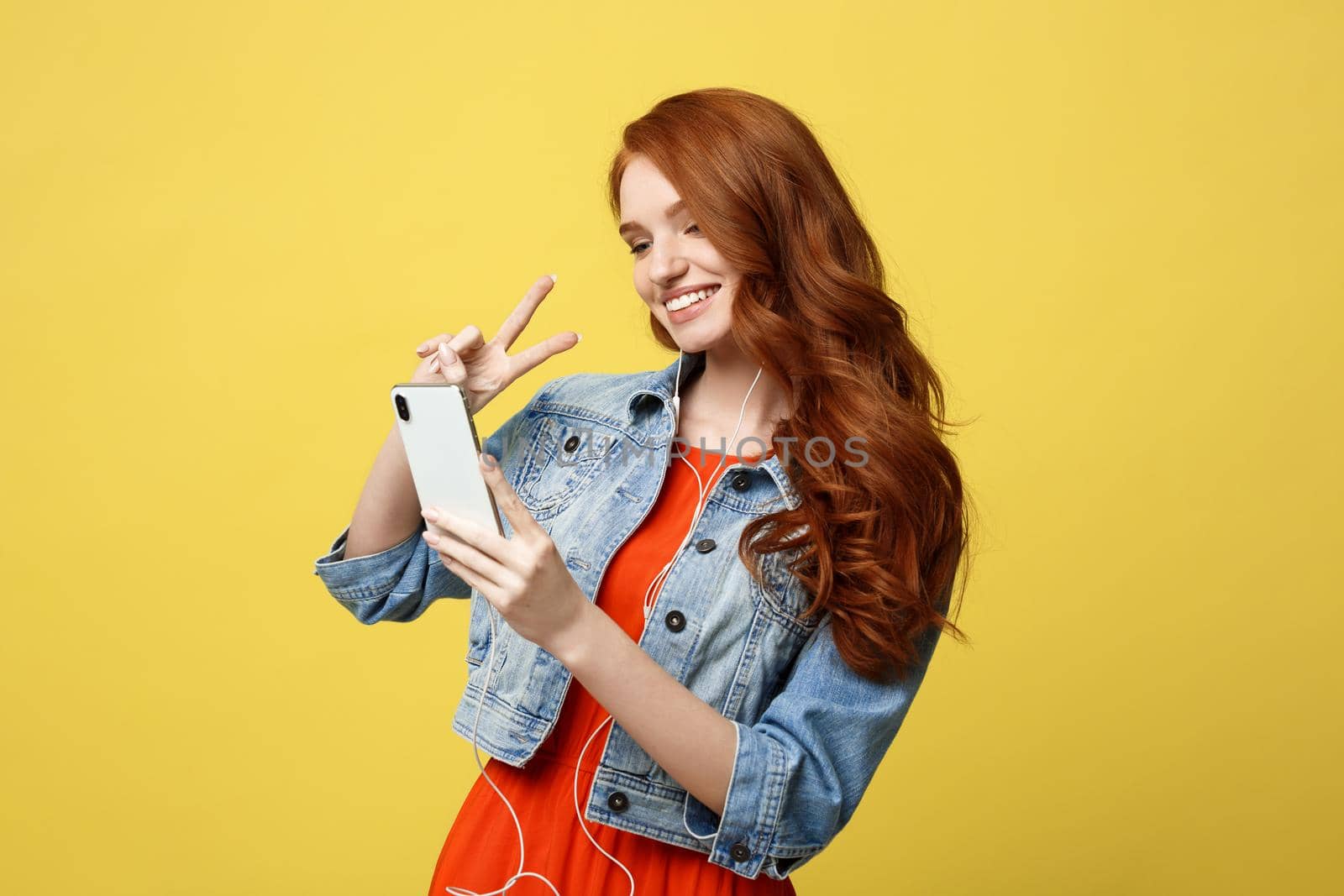 Lifestyle Concept - Portrait ginger hair woman in casual dress using mobile phone for interaction video calling by showing two fingers. Isolated on bright yellow background.