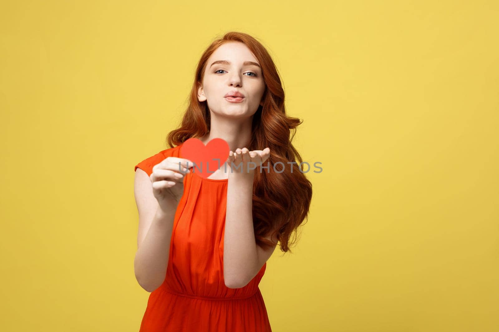 Valentine day, love and romance concept: Happy girl blowing a kiss. Young woman in bright casual clothes with paper heart at yellow background