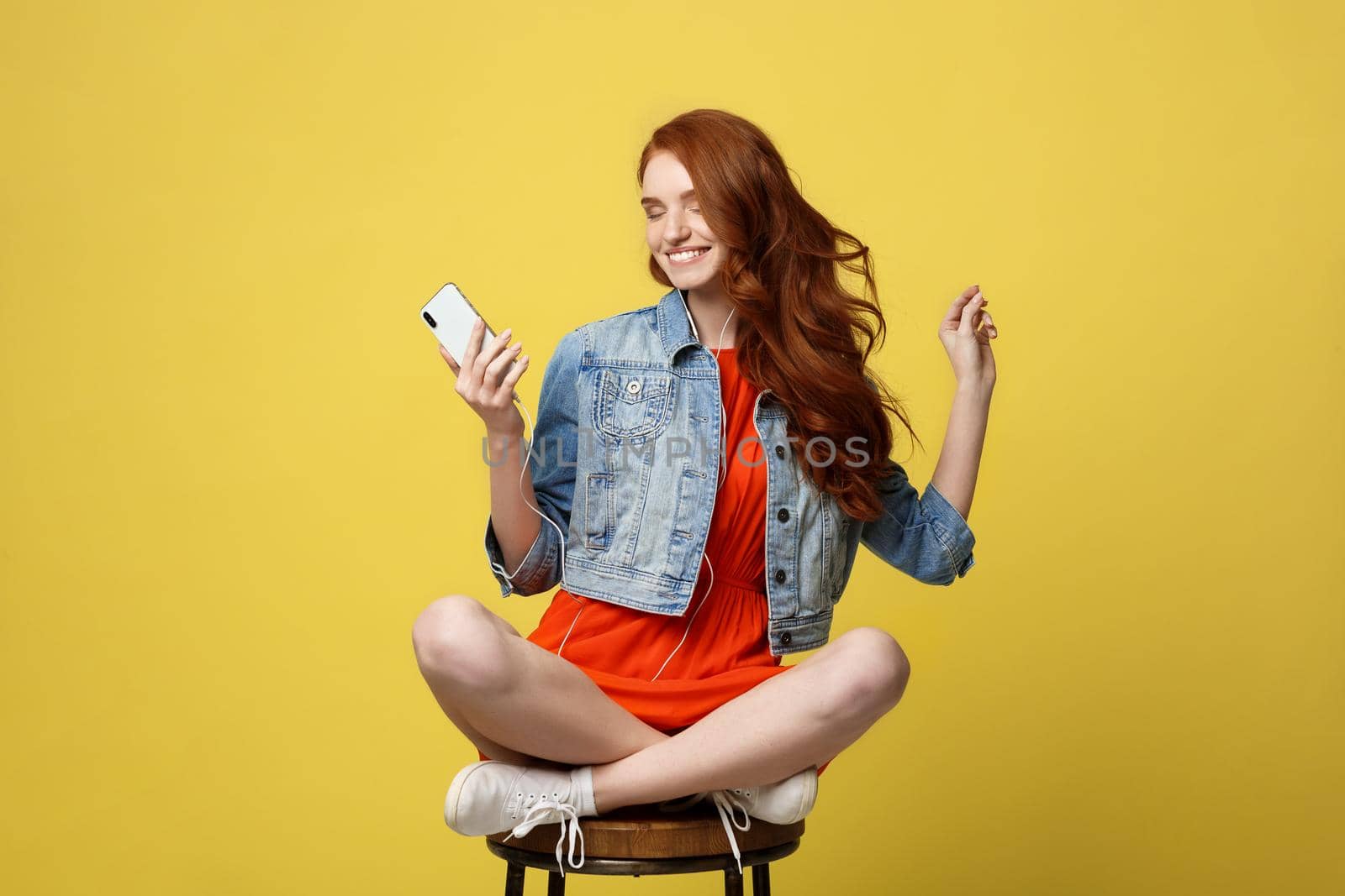 Lifestyle, Music, Technology concept: Young beautiful caucasian woman listening music with headphones and smart phone hand hold, dancing, eyes closed smiling, Isolated on yellow vivid background