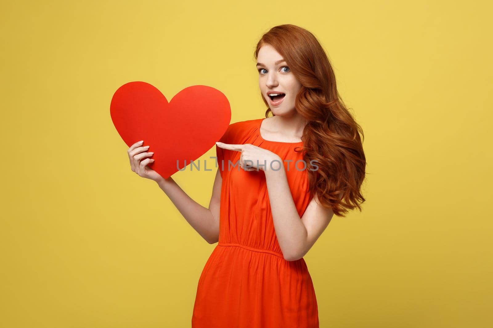 Valentine and Lifestyle Concept: Portrait of an attractive young woman dressed in red dress pointing finger at paper heart isolated over yellow studio background