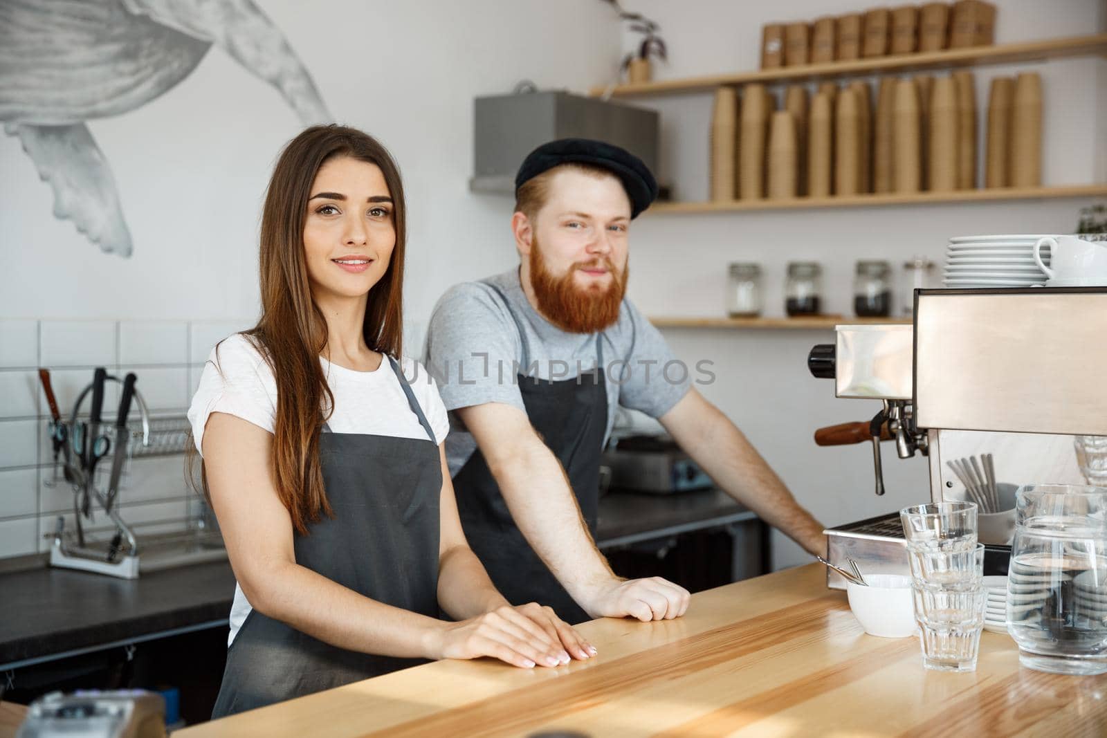 Coffee Business Concept - Positive young bearded man and beautiful attractive lady barista couple in apron while standing at bar Couter ready to give Coffee Service at the modern coffee shop by Benzoix