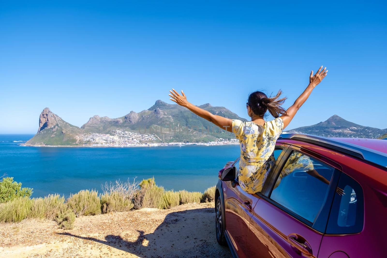 woman outside car window road trip with hands up, car at Chapmans Peak Drive in Cape Town South Africa looking out over the ocean.