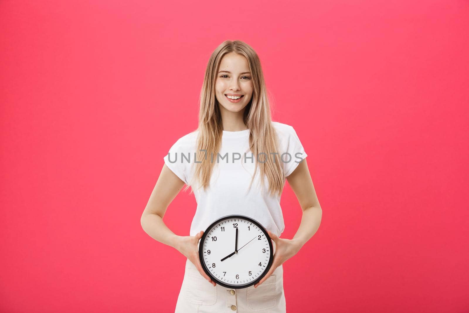 Portrait of an excited young girl dressed in white t-shirt pointing at alarm clock and looking at camera isolated over pink background by Benzoix