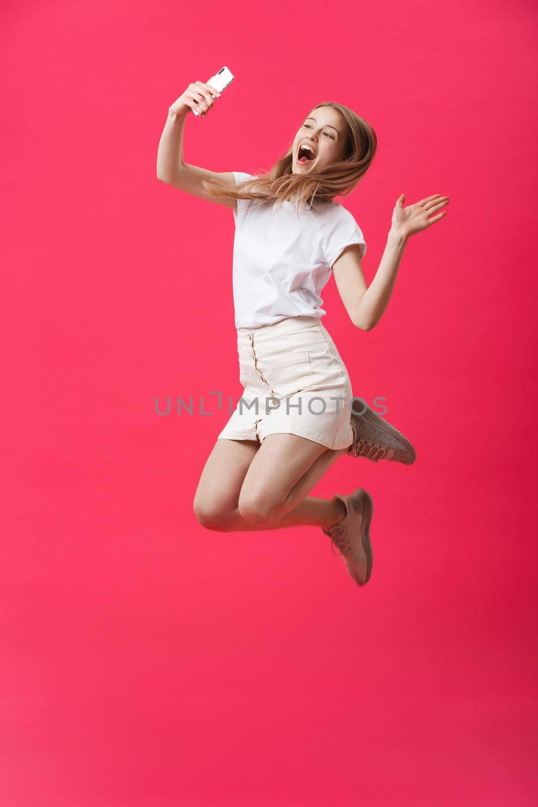 Full length portrait of an excited teenage schoolgirl in uniform with taking a selfie while jumping and showing say hi gesture isolated over pink background