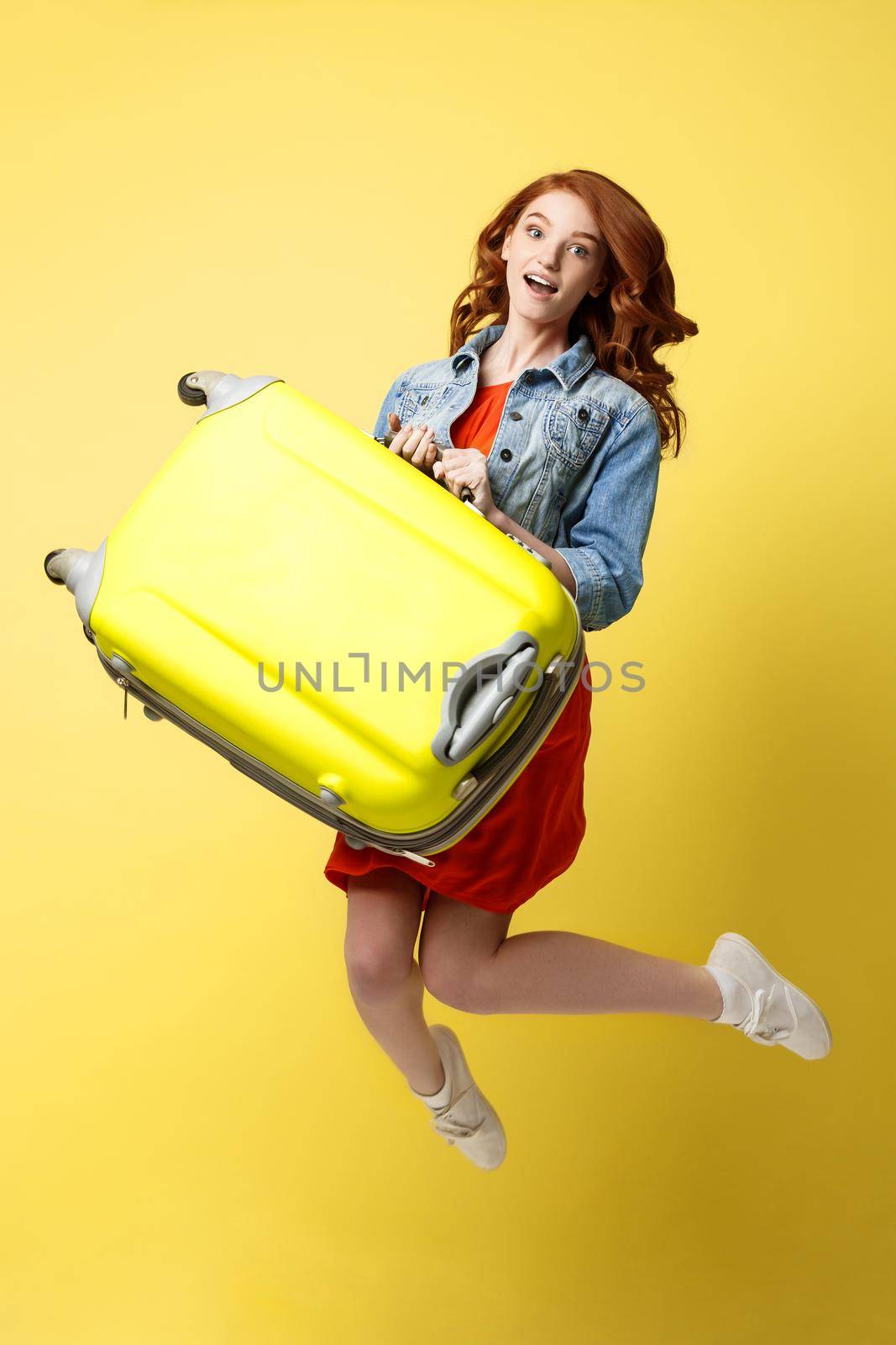 Travel and Lifestyle Concept: Portrait of a beautiful red hair fashion woman jumping and holding a bright green suitcase isolated over bright yellow background. by Benzoix