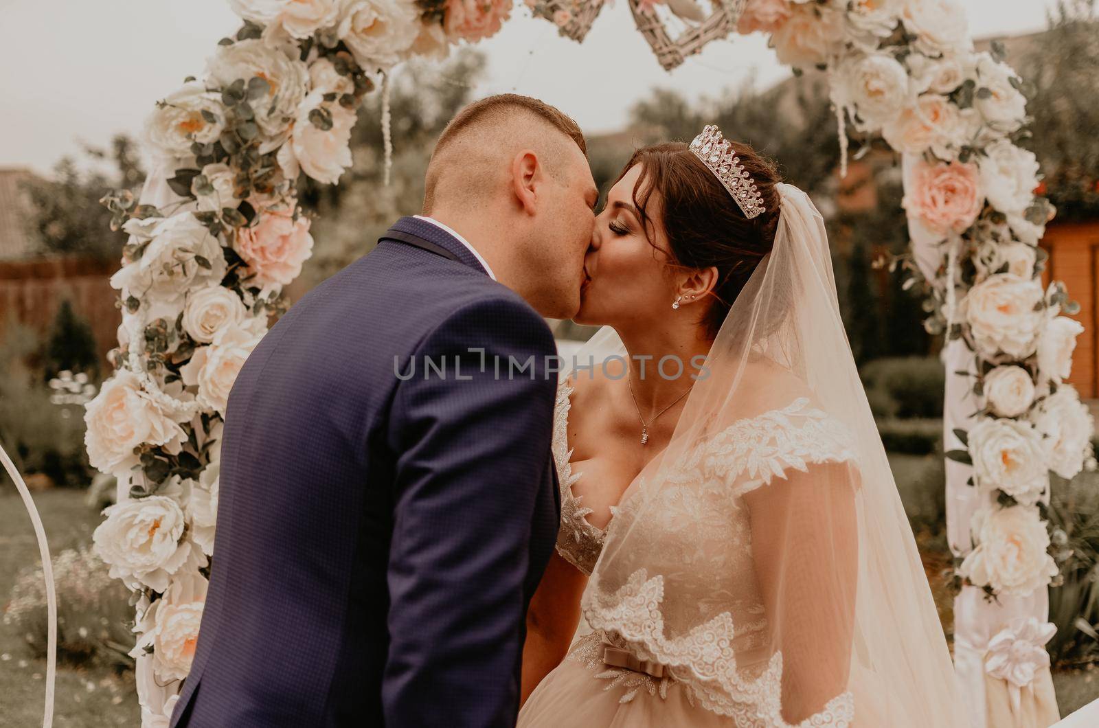 Bride and groom in a wedding dress and a long veil at a wedding ceremony on the background of a flower arch laughing kissing rejoicing. Slavic Ukrainian Russian traditions