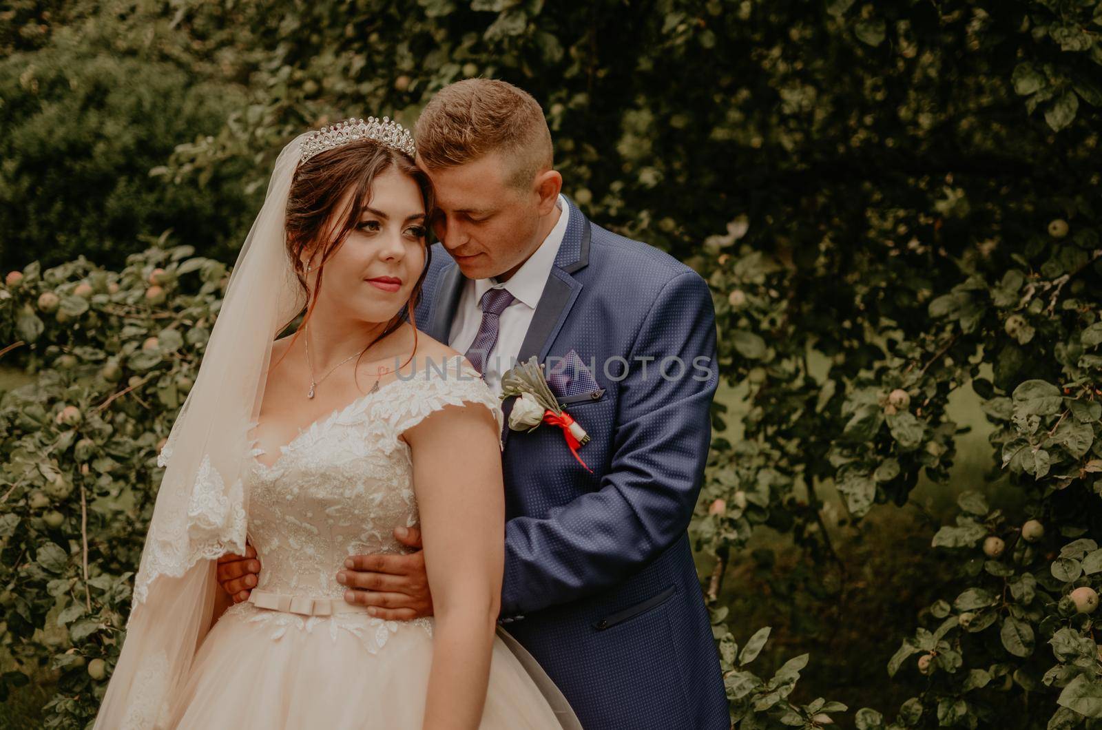 blonde European Caucasian young man groom in blue suit and black-haired woman bride in white wedding dress with long veil and tiara on head. newlyweds guy hugs a girl from behind