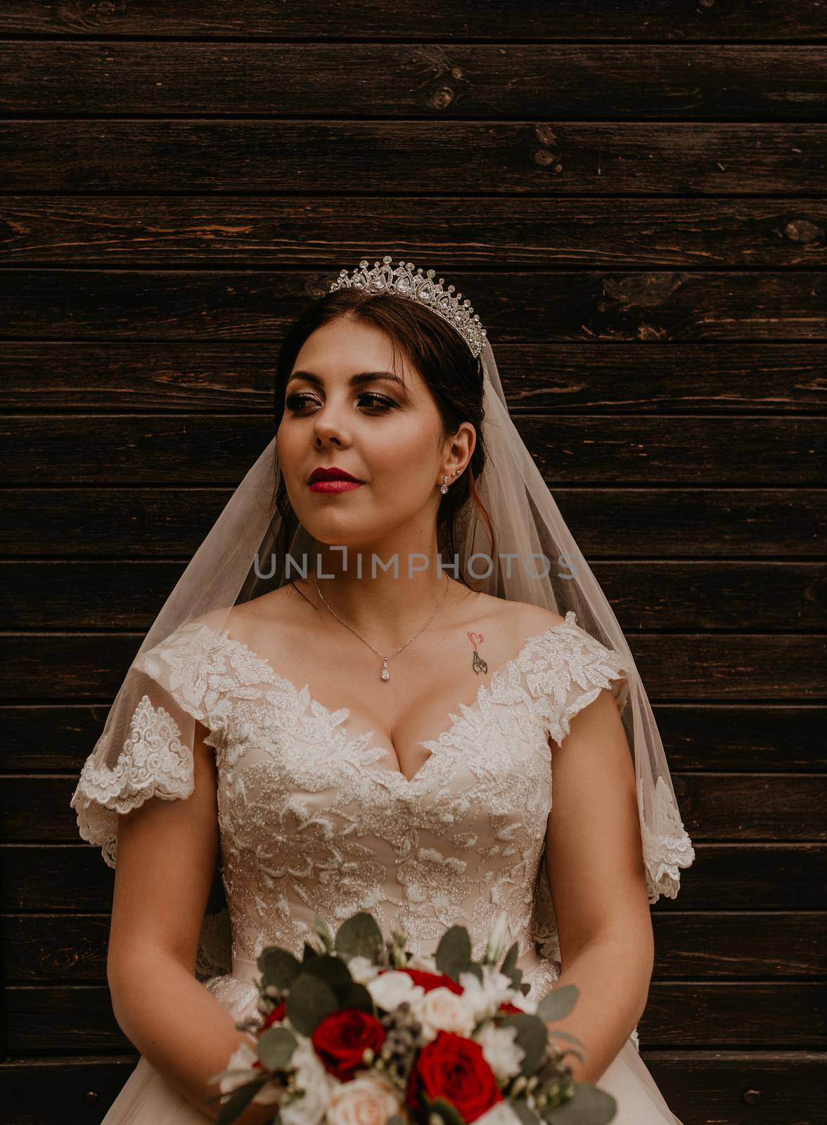 European Caucasian young black-haired woman bride in white wedding dress with long veil and tiara on head. girl holding her bouquet of flowers in hands. dark brown wooden background with planks