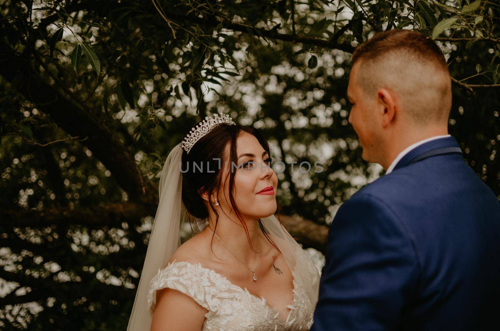 blonde European Caucasian young man groom in blue suit and black-haired woman bride in white wedding dress with long veil and tiara on head Newlyweds under a tree in summer