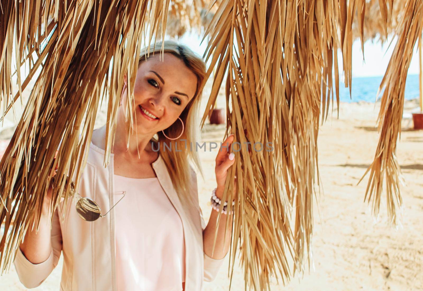 Beautiful blonde woman on a background of dry leaves and palm branches on the beach. Blonde girl wearing pink clothes Against the background of palm trees and the sea. Enjoys vacation in Egypt.