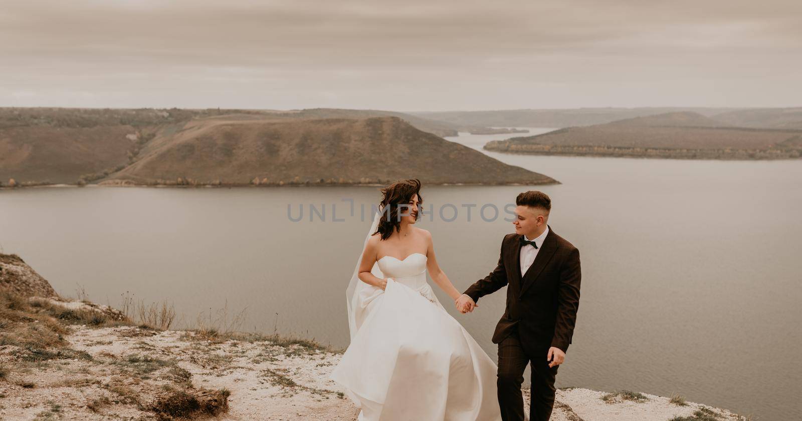 bride in white dress long veil and groom in suit walk in summer fall on mountain above river by AndriiDrachuk