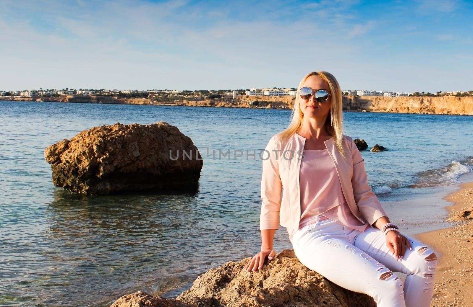 Blonde girl in sunglasses sits on a stone near the sea. by ViShark