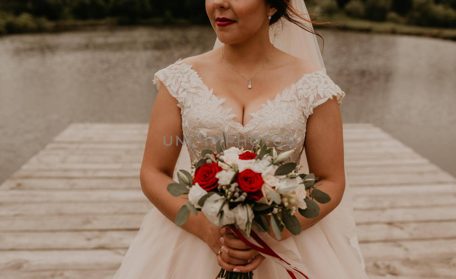 European Caucasian young black-haired woman bride in white wedding dress with long veil and tiara on head. girl holding her bouquet flowers in hands. bride stands on wooden pier on background of river