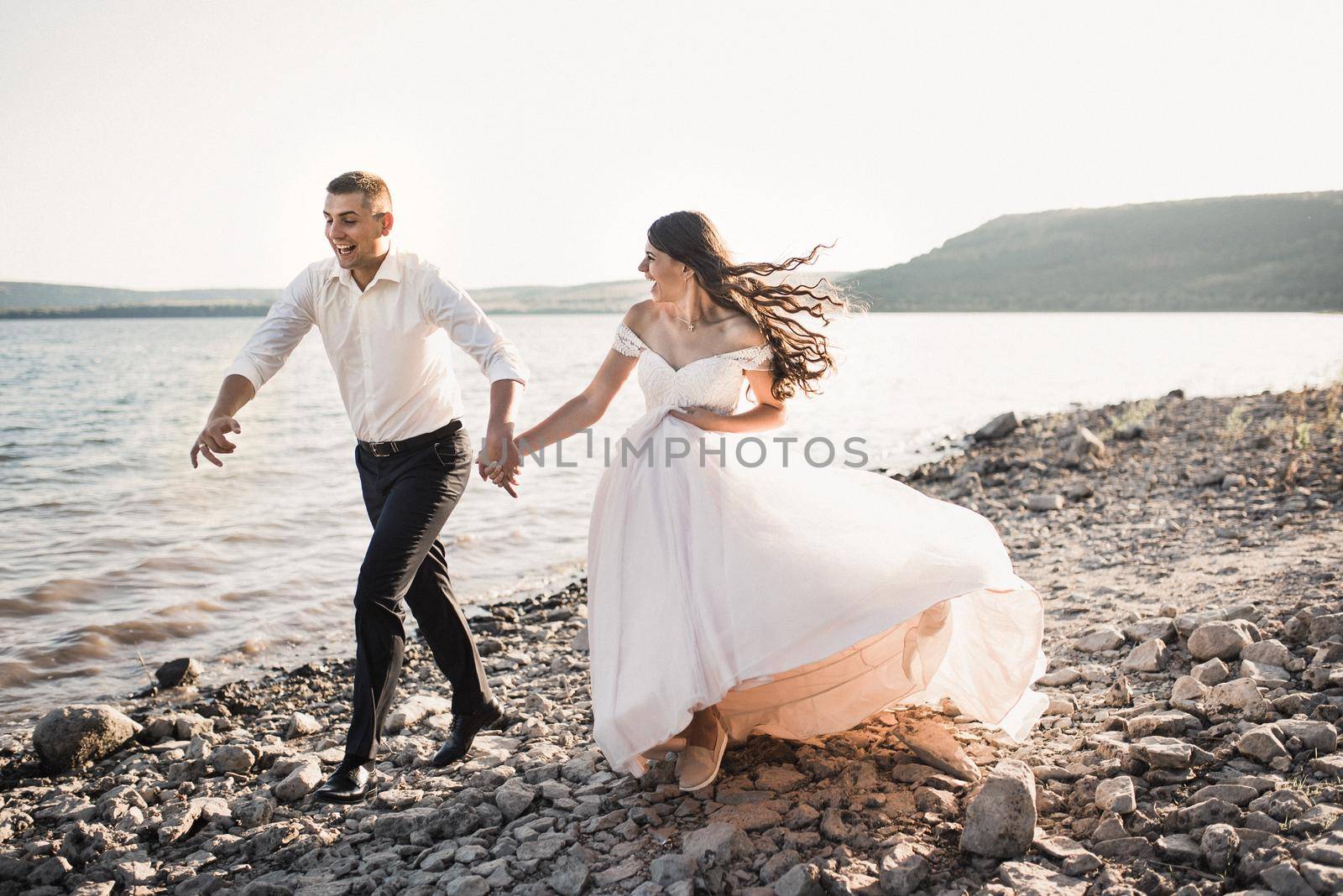 bride and groom laughing run together against rays of setting sun in summer by AndriiDrachuk