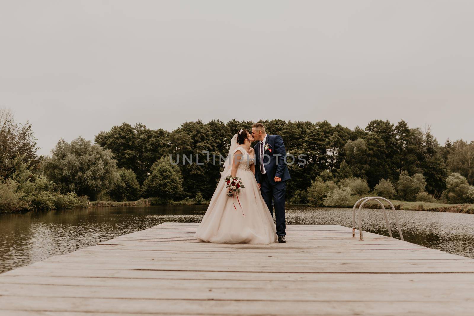 Newlyweds kissing hugging on wooden pier bridge on lake by AndriiDrachuk