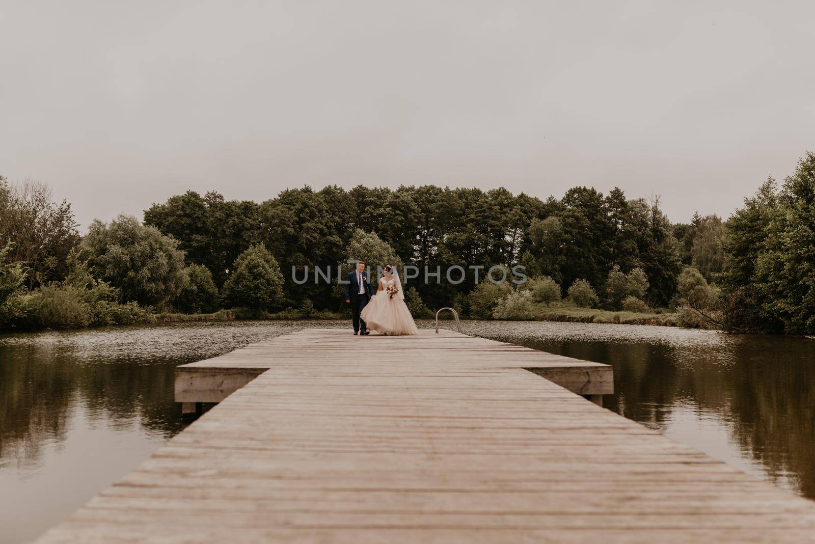 blonde European Caucasian young man groom in blue suit and black-haired woman bride in white wedding dress with long veil and tiara on head. Newlyweds kissing hugging on wooden pier bridge on lake