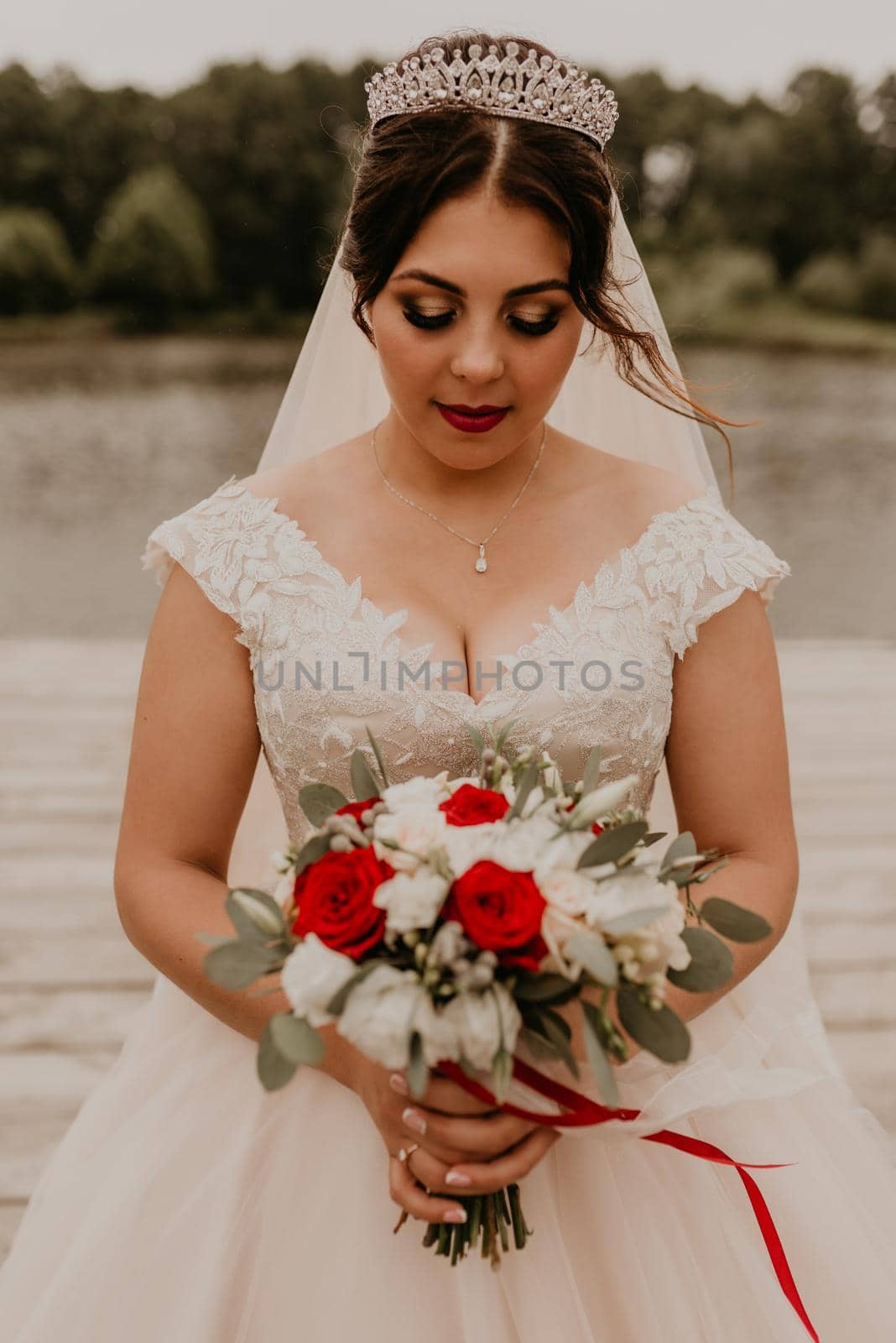 European Caucasian young black-haired woman bride in white wedding dress with long veil and tiara on head. girl holding her bouquet flowers in hands. bride stands on wooden pier on background of river