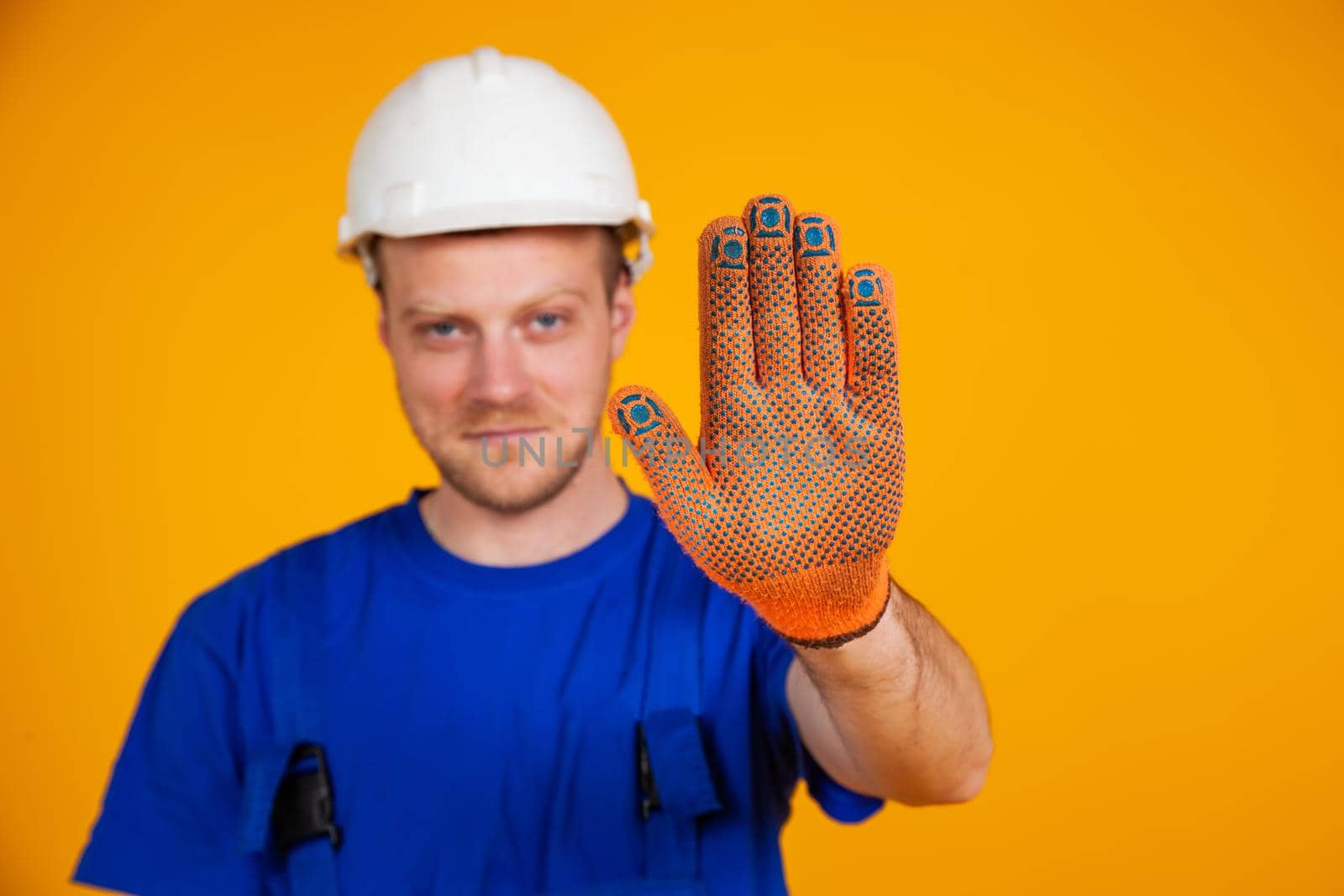 Male worker shows stop hand gesture. Worker in overalls and protective helmet shows stop gesture with gloved hand.