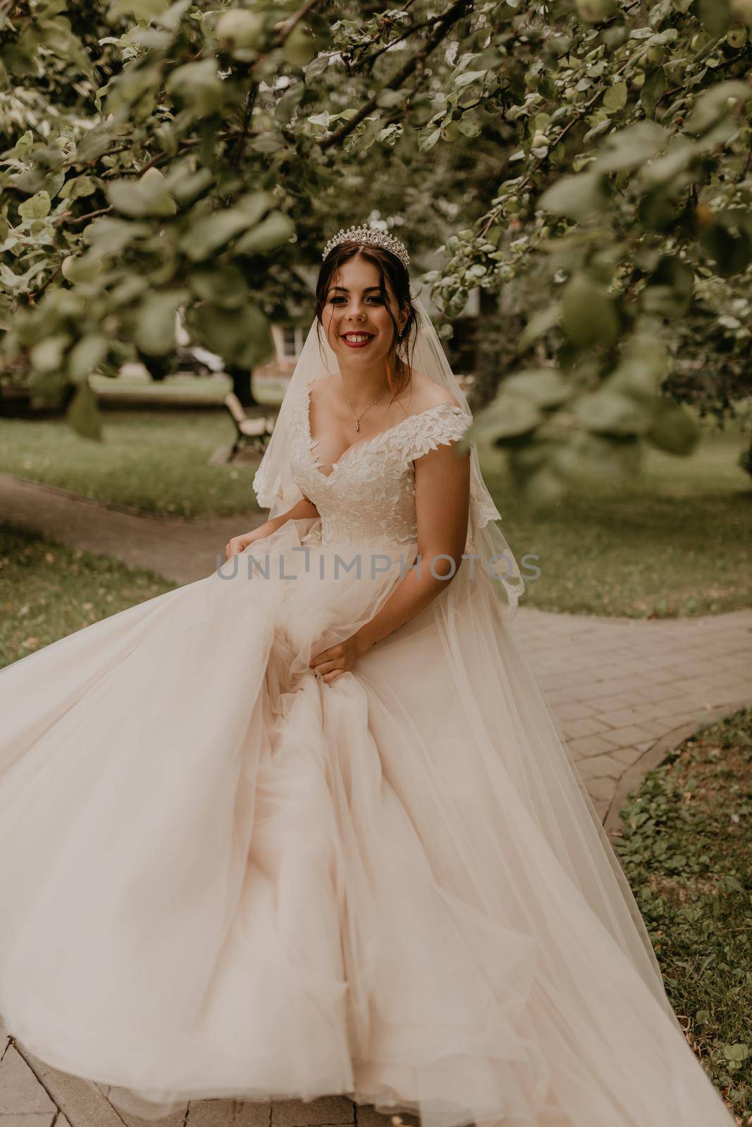 dark-haired European Caucasian young woman bride in white wedding dress with long veil and tiara on head spinning whirls dancing posing moving at nature on background greenery. Valentine love couple