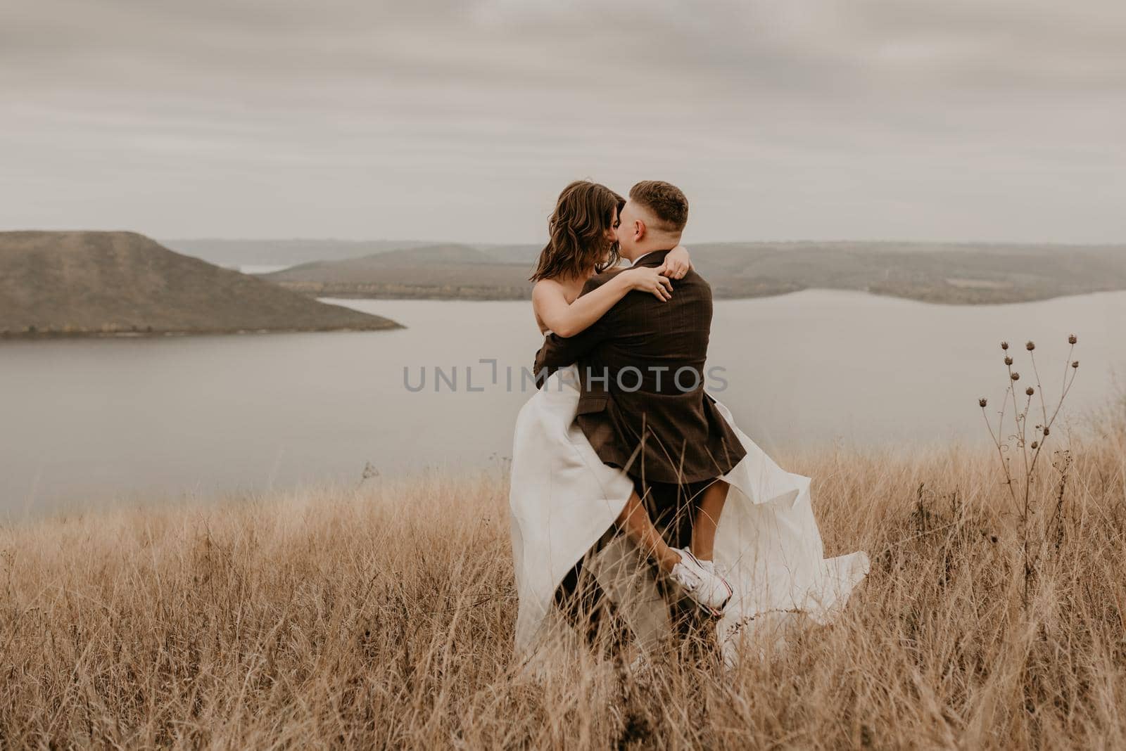 loving couple wedding newlyweds in dress veil sports shoes and suit hug kissing whirl on tall grass in field on mountain above river by AndriiDrachuk