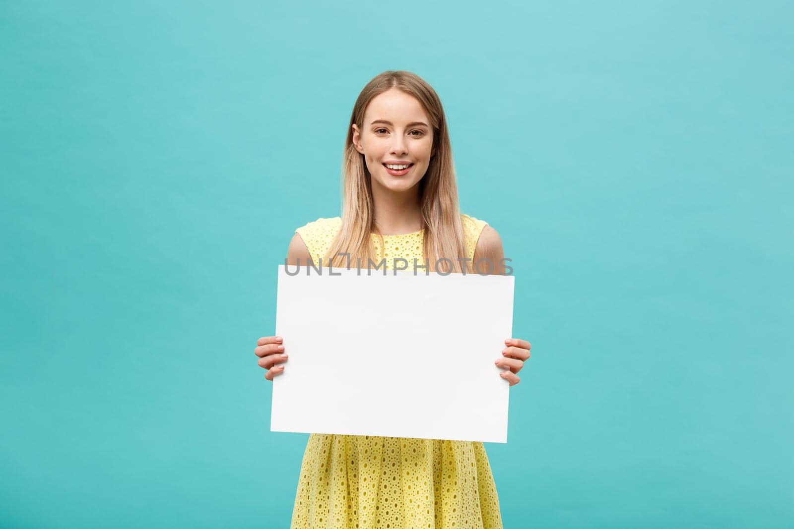 Lifestyle Concept: young beautiful girl smiling and holding a blank sheet of paper, dressed in yellow, isolated on pastel blue background by Benzoix