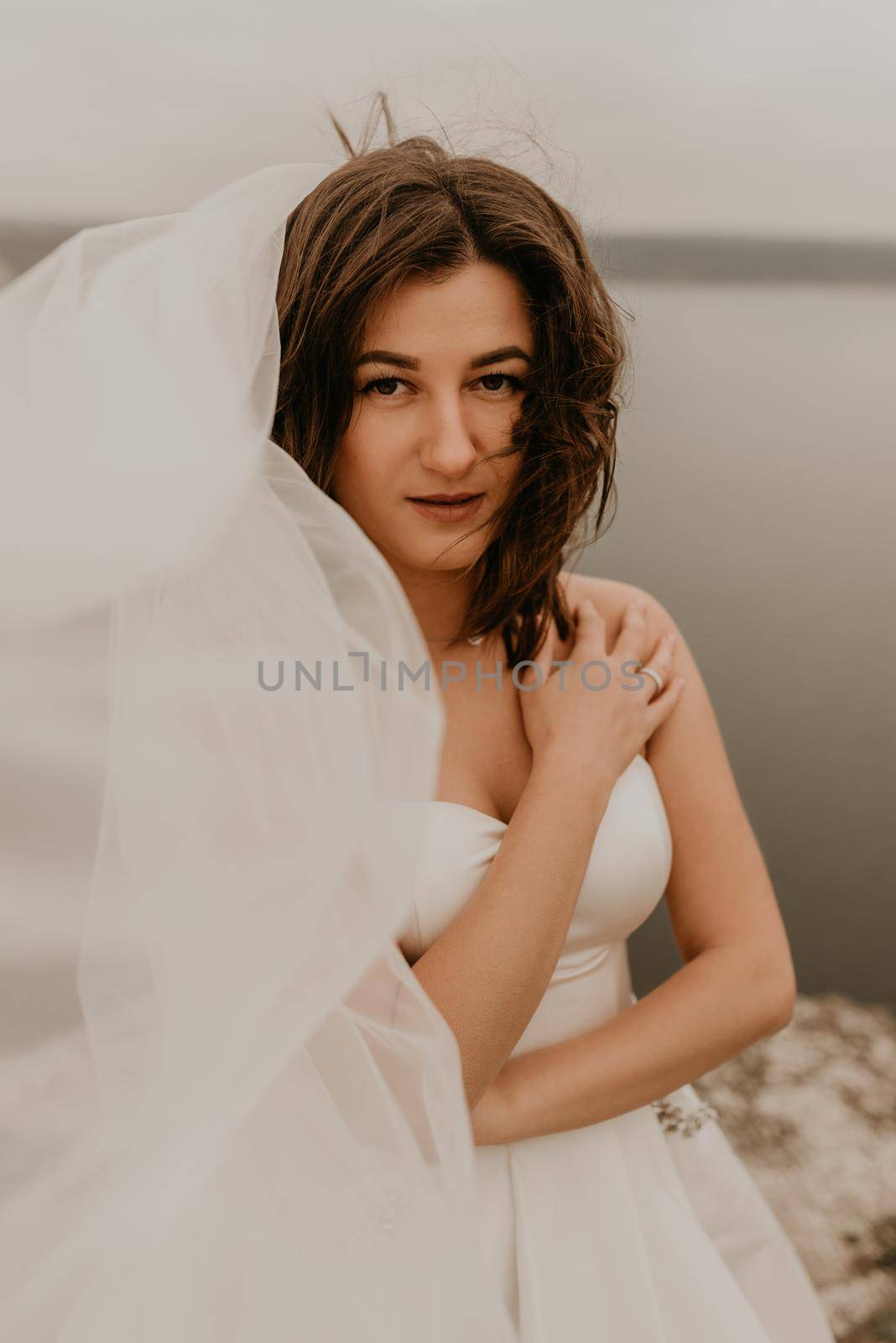 young brunette bride woman with short haircut in white wedding corset dress strapless and with a long chiffon veil on head. natural light nude makeup. Cool autumn weather. background large lake river