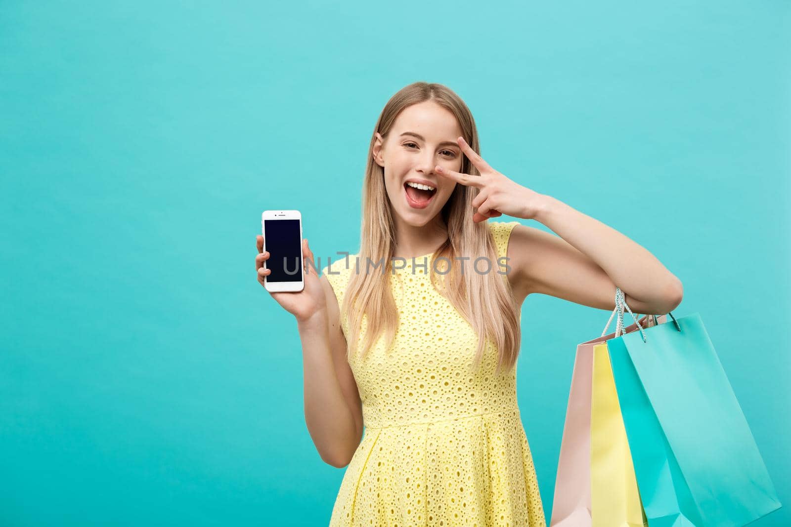 Portrait young attractive woman with shopping bags shows the phone's screen directly to the camera. Isolated on blue background.