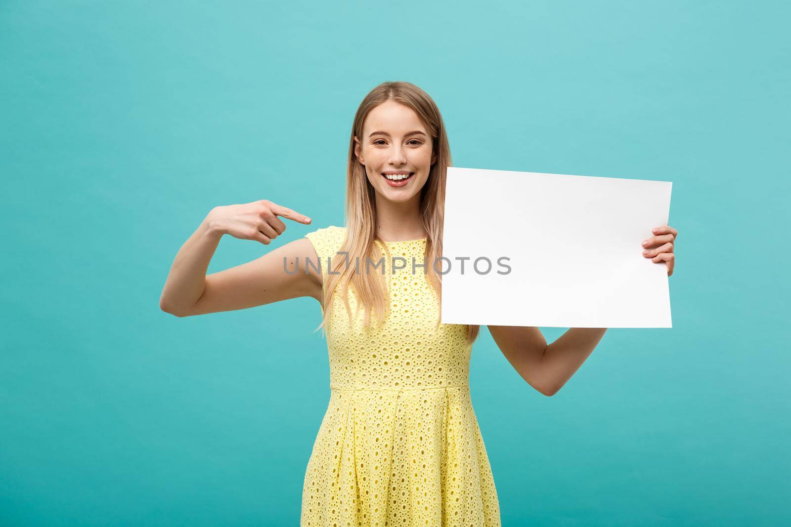Portrait of young woman in yellow dress pointing finger at side white blank board. Isolated over Blue background
