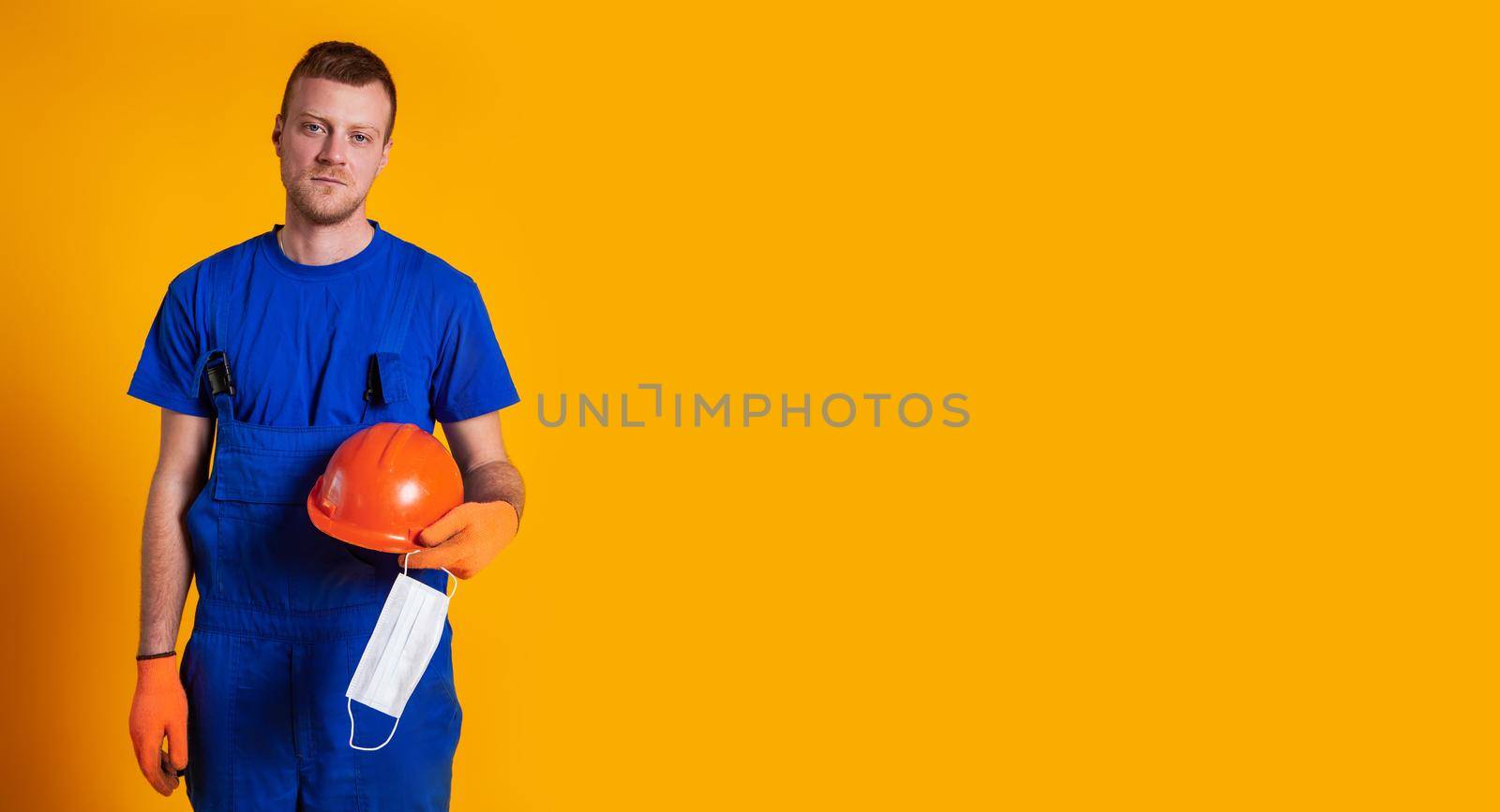 Worker in construction overalls and gloves on a yellow background, holds a protective helmet and a medical mask in his hand. The concept of the economic crisis, unemployment production, coronavirus.