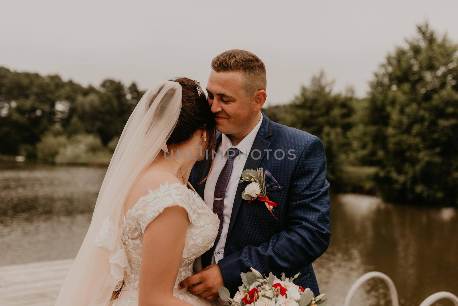 blonde European Caucasian young man groom in blue suit and black-haired woman bride in white wedding dress with long veil and tiara on head. Newlyweds kissing hugging on wooden pier bridge on lake