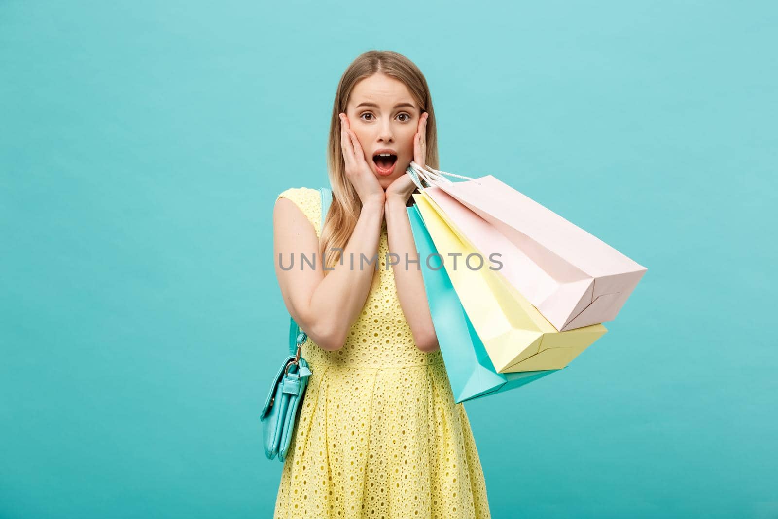 Lifestyle Concept: Portrait shocked young brunette woman in yellow summer dress posing with shopping bags isolated over pastel blue background.