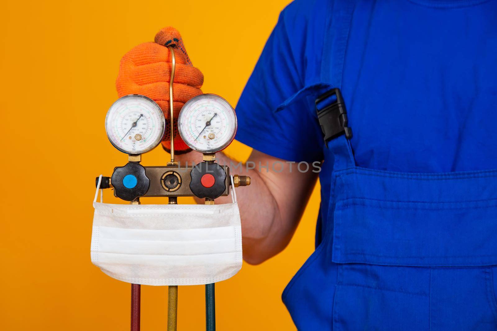 An air conditioning technician holds pressure gauges in his hand for refueling air conditioners. Medical face shield on measuring equipment for filling air conditioners.