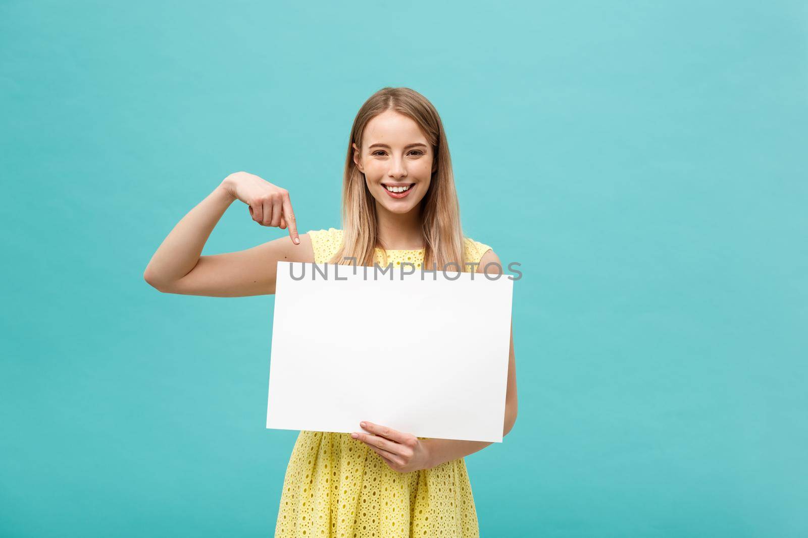 Portrait of young woman in yellow dress pointing finger at side white blank board. Isolated over Blue background. by Benzoix