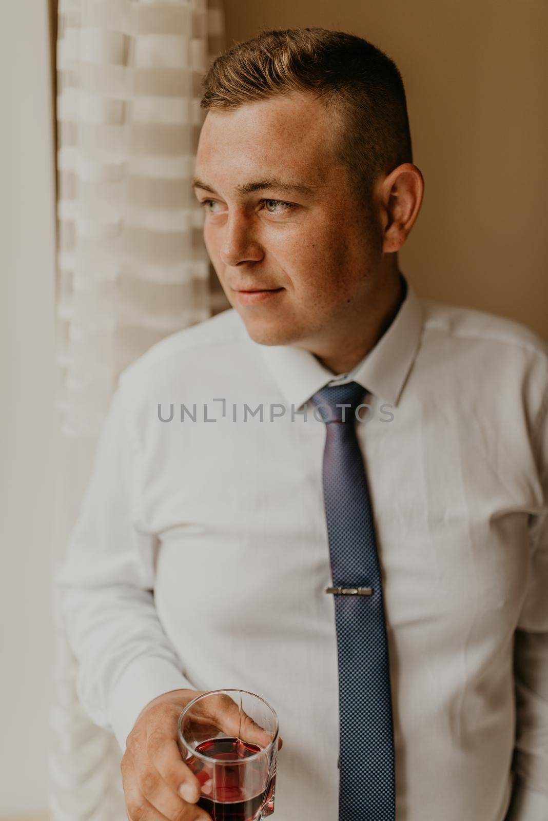 blonde European Caucasian young groom in shirt business suit with tie prepare for wedding. preparing for important event meeting indoors worries. a man holding a glass of whiskey in his hands drinking