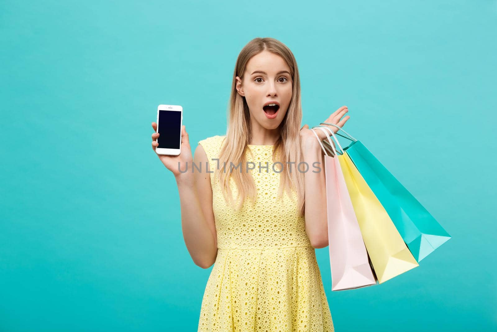 Portrait young attractive woman with shopping bags shows the phone's screen directly to the camera. Isolated on blue background.
