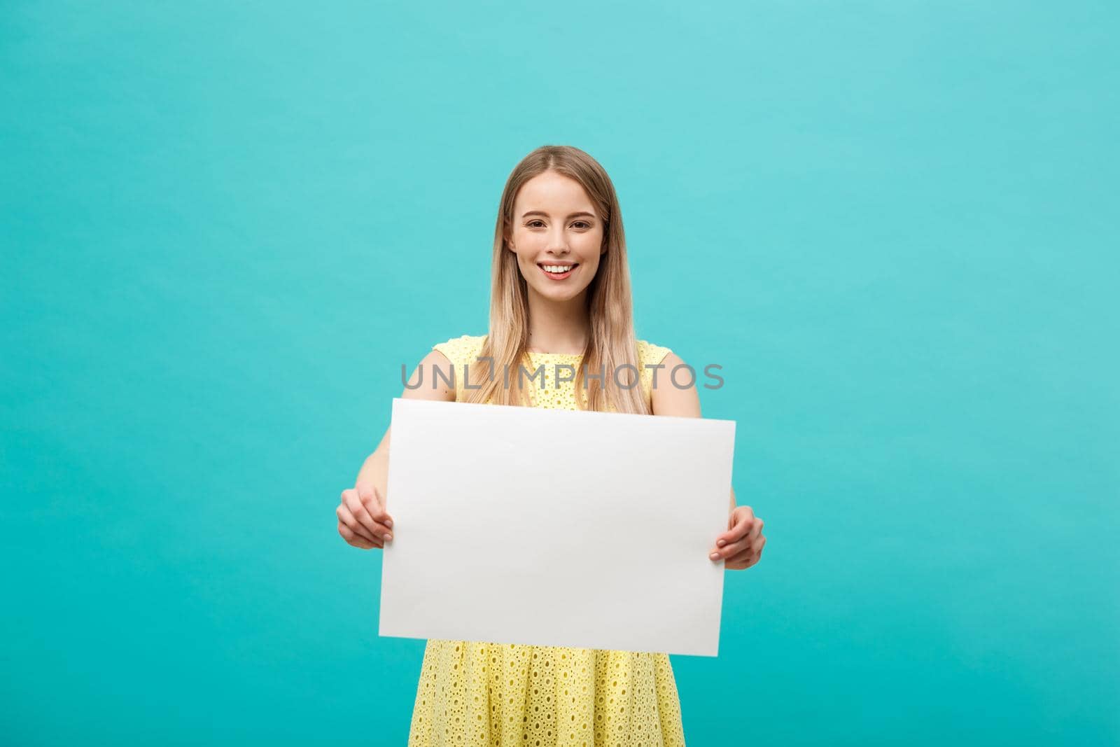 Lifestyle Concept: young beautiful girl smiling and holding a blank sheet of paper, dressed in yellow, isolated on pastel blue background.