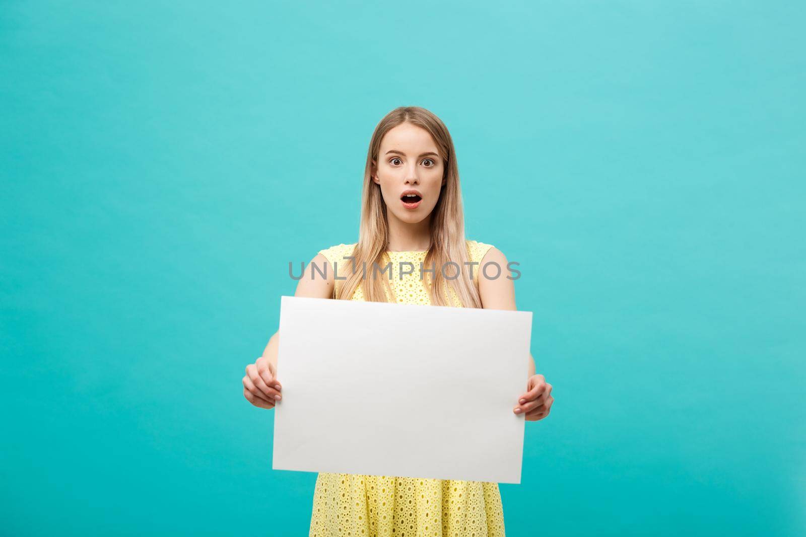 Portrait of amazed young blond woman holding blank sign with copy space on blue studio background. Showing shocked surprise face. by Benzoix