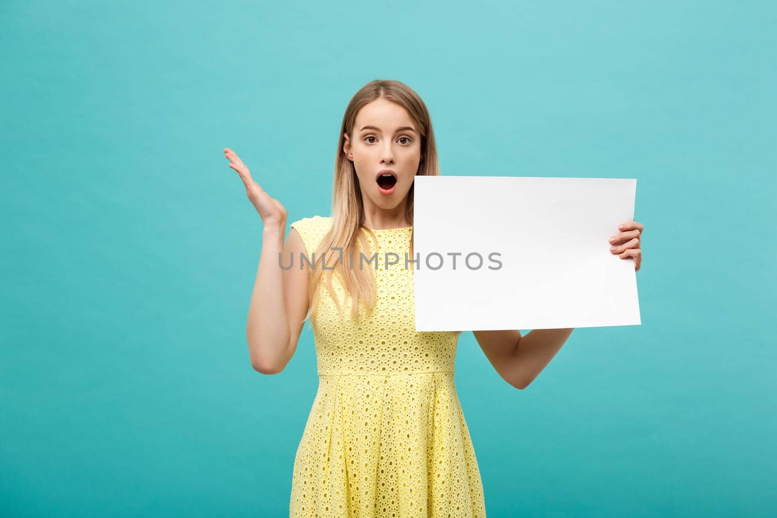 Closeup portrait, young woman in yellow dress holding white plain paper with shocked expression at what she sees, isolated blue background