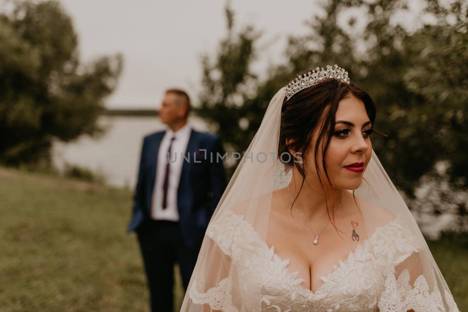 European Caucasian young man groom in blue suit and black-haired woman bride in white wedding dress with long veil and tiara on head. newlyweds look in different directions. man stands behind woman