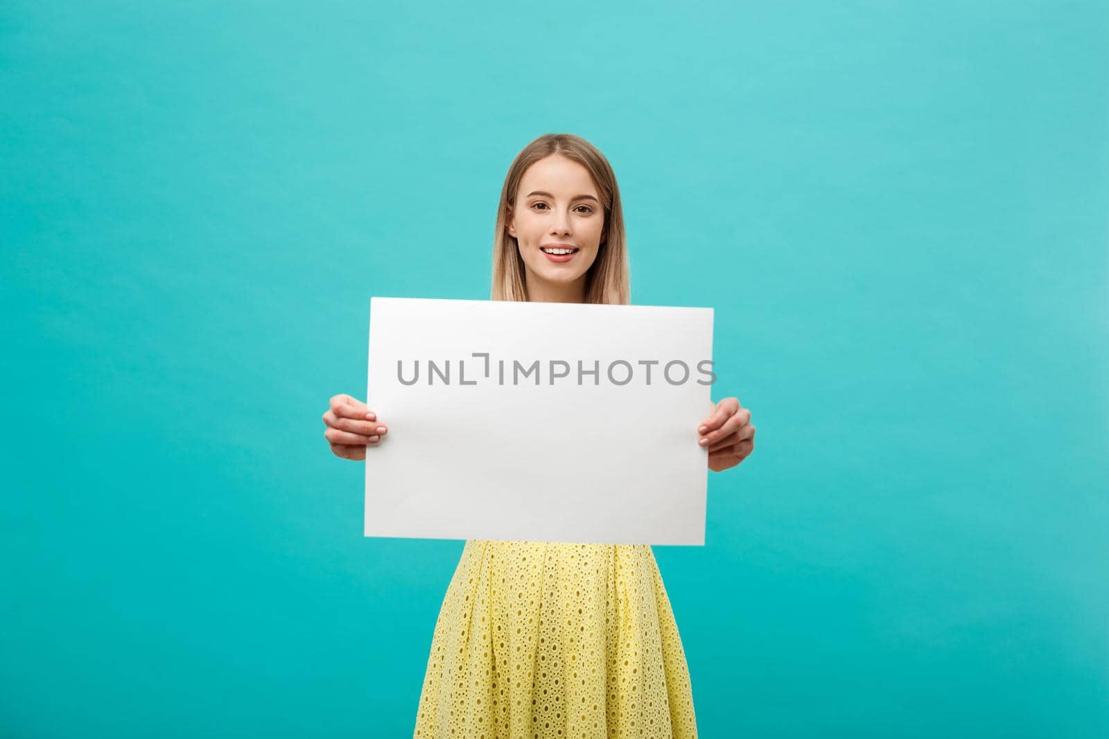 Lifestyle Concept: young beautiful girl smiling and holding a blank sheet of paper, dressed in yellow, isolated on pastel blue background.