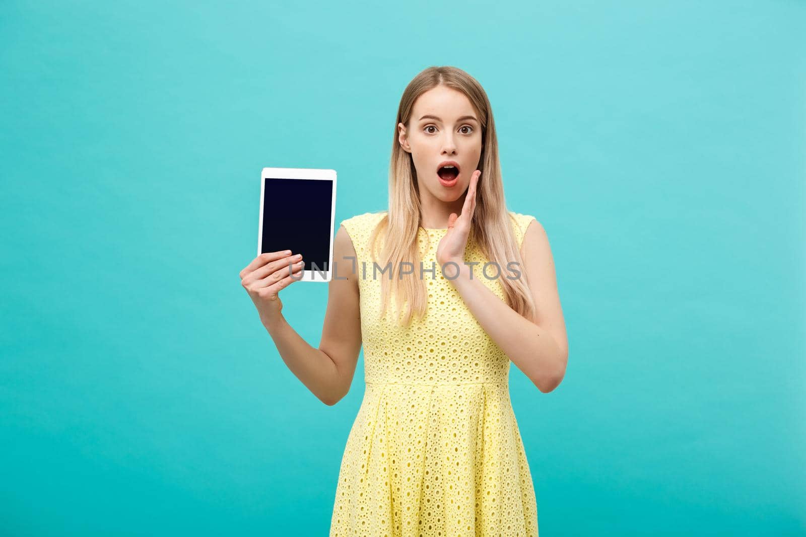 Portrait of a surprised amazed young woman looking at digital tablet isolated on a blue studio background. by Benzoix