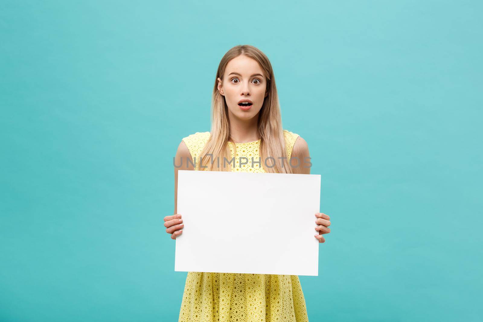 Portrait of amazed young blond woman holding blank sign with copy space on blue studio background. Showing shocked surprise face