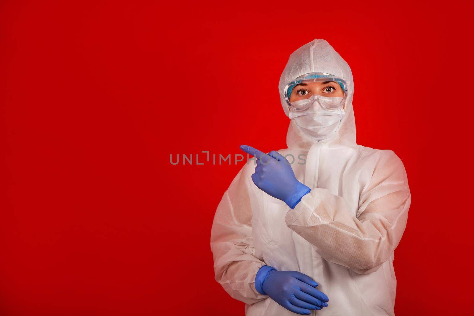 A woman in a protective suit against the COVID-19 virus indicates the direction. Medic woman wearing protective clothing against coronovirus on a red background.