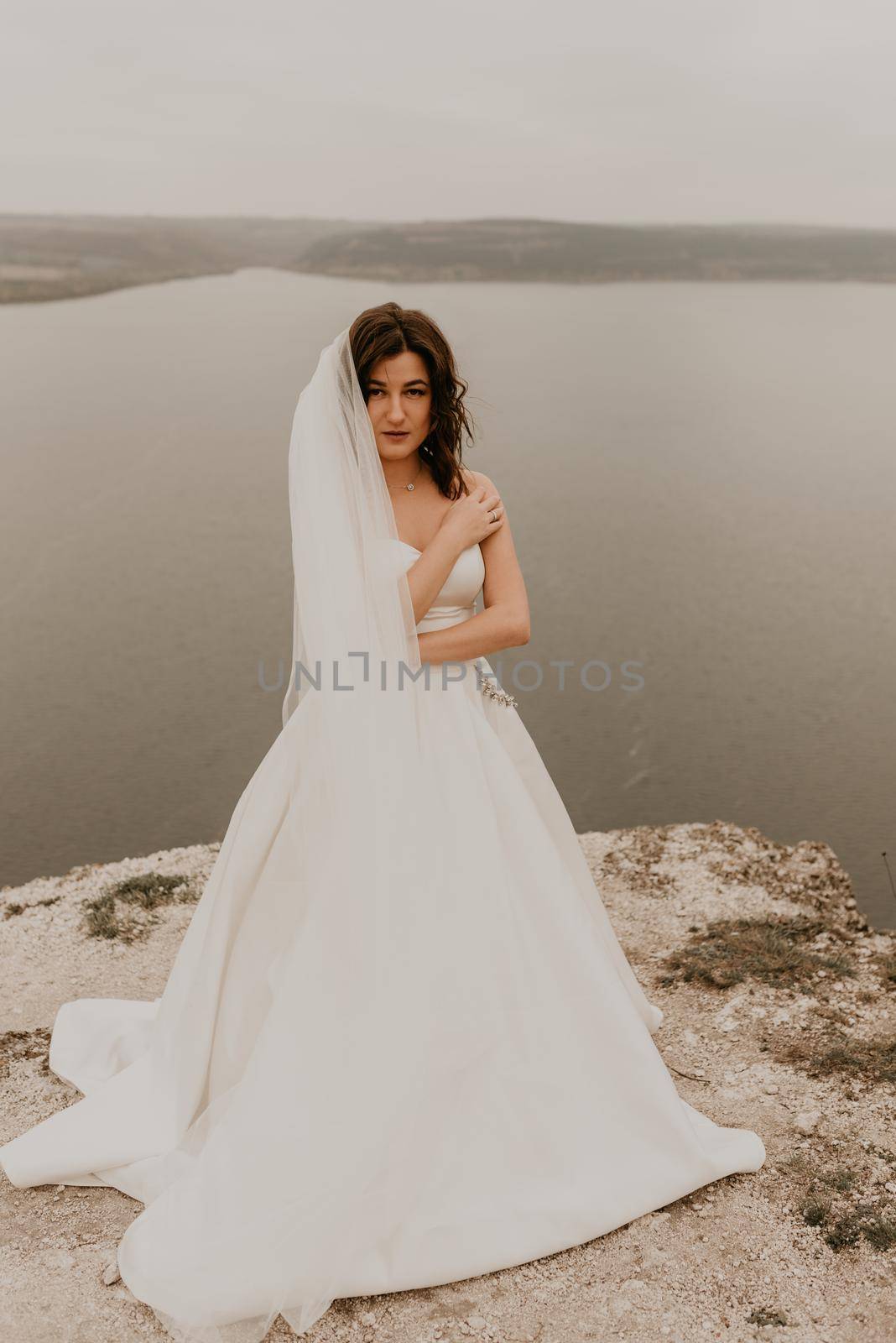 young brunette bride woman with short haircut in white wedding corset dress strapless and with a long chiffon veil on head. natural light nude makeup. Cool autumn weather. background large lake river