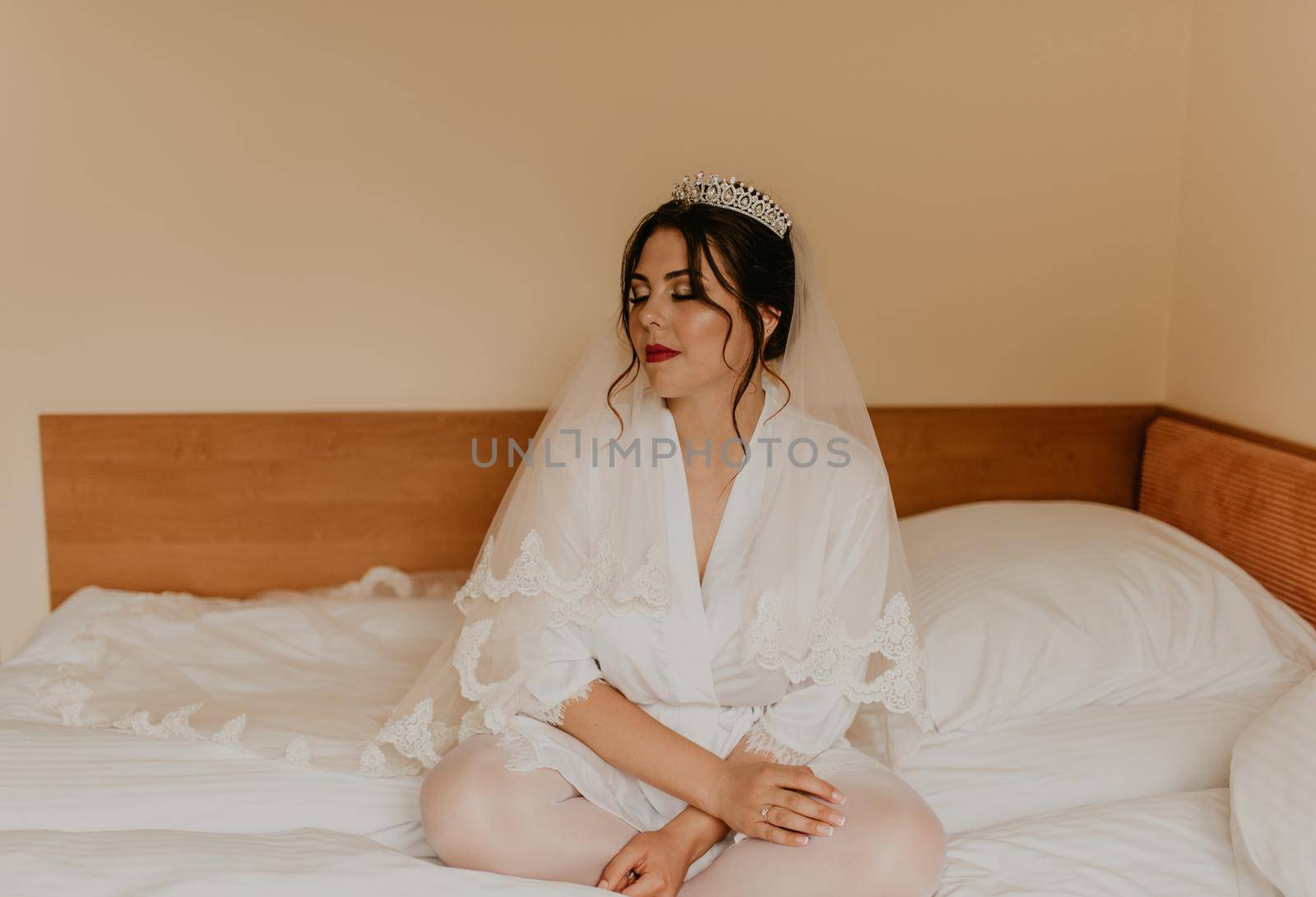 European Caucasian young black-haired woman bride white silk peignoir thin robe long veil tiara on head. morning gatherings of preparing bride for holiday party wedding. girl sitting on bed in bedroom