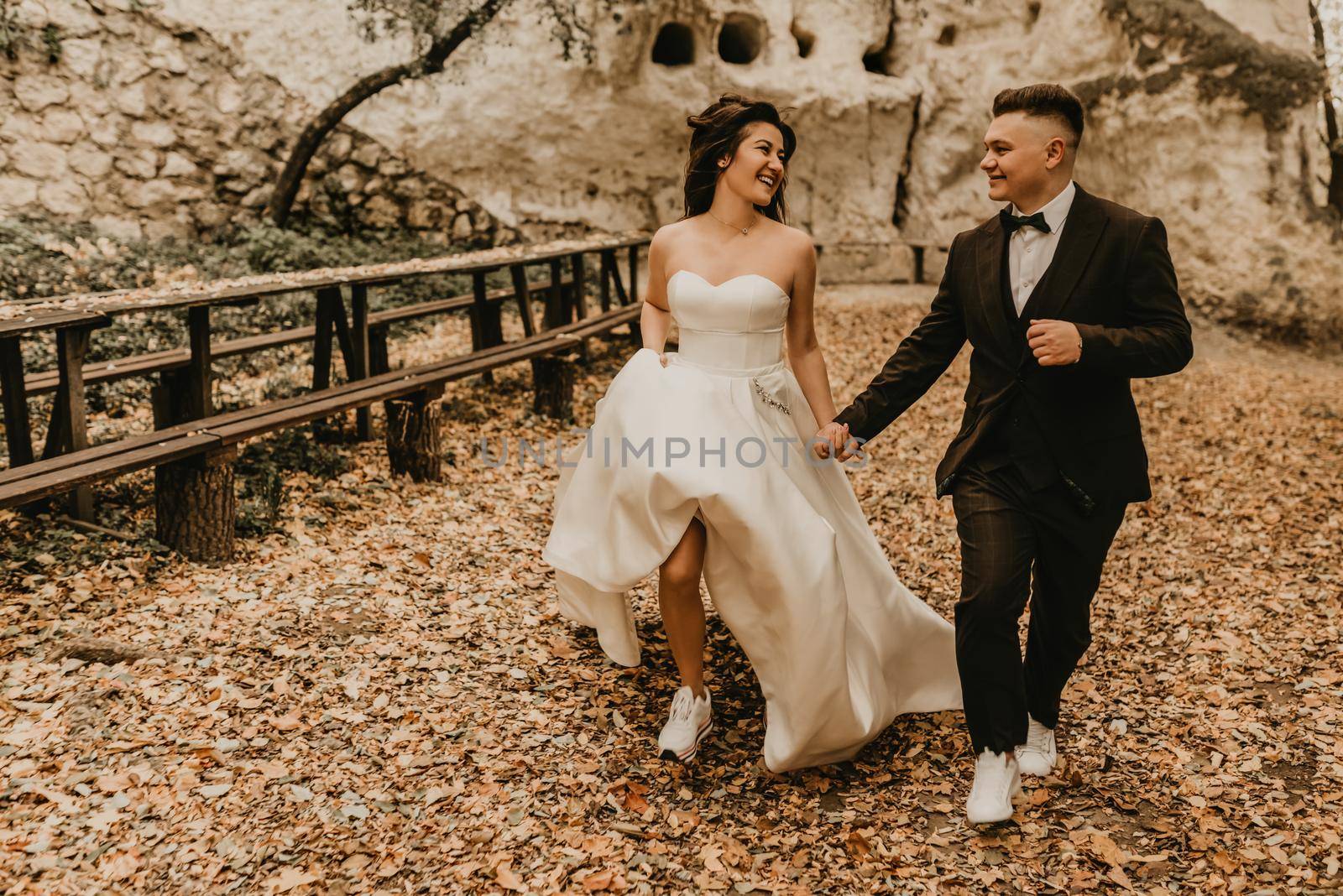wedding couple in love man and woman run happy autumn forest background of stone rocks. groom in suit and bride dress with long veil on short hair sports shoes outdoors. rock monastery in bakota
