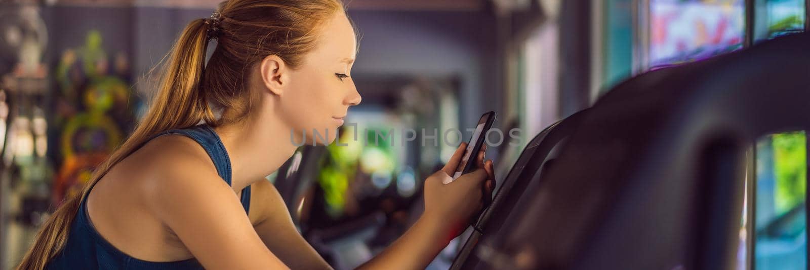 Young woman using phone while training at the gym. Woman sitting on exercising machine holding mobile phone BANNER, LONG FORMAT by galitskaya