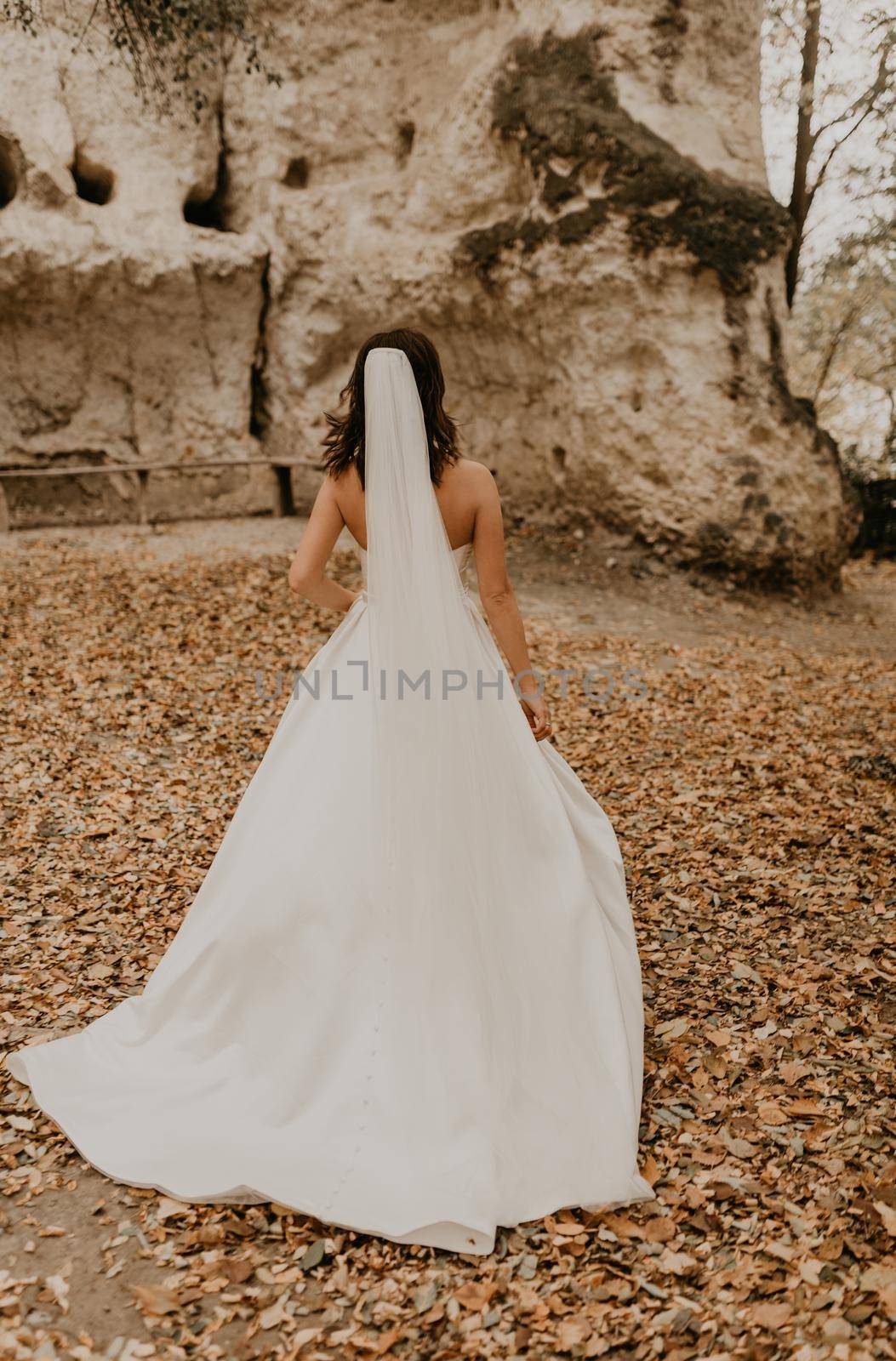young woman bride in white wedding dress with long veil run away through autumn forest on fallen orange leaves to mountain. brunette with short hair bob