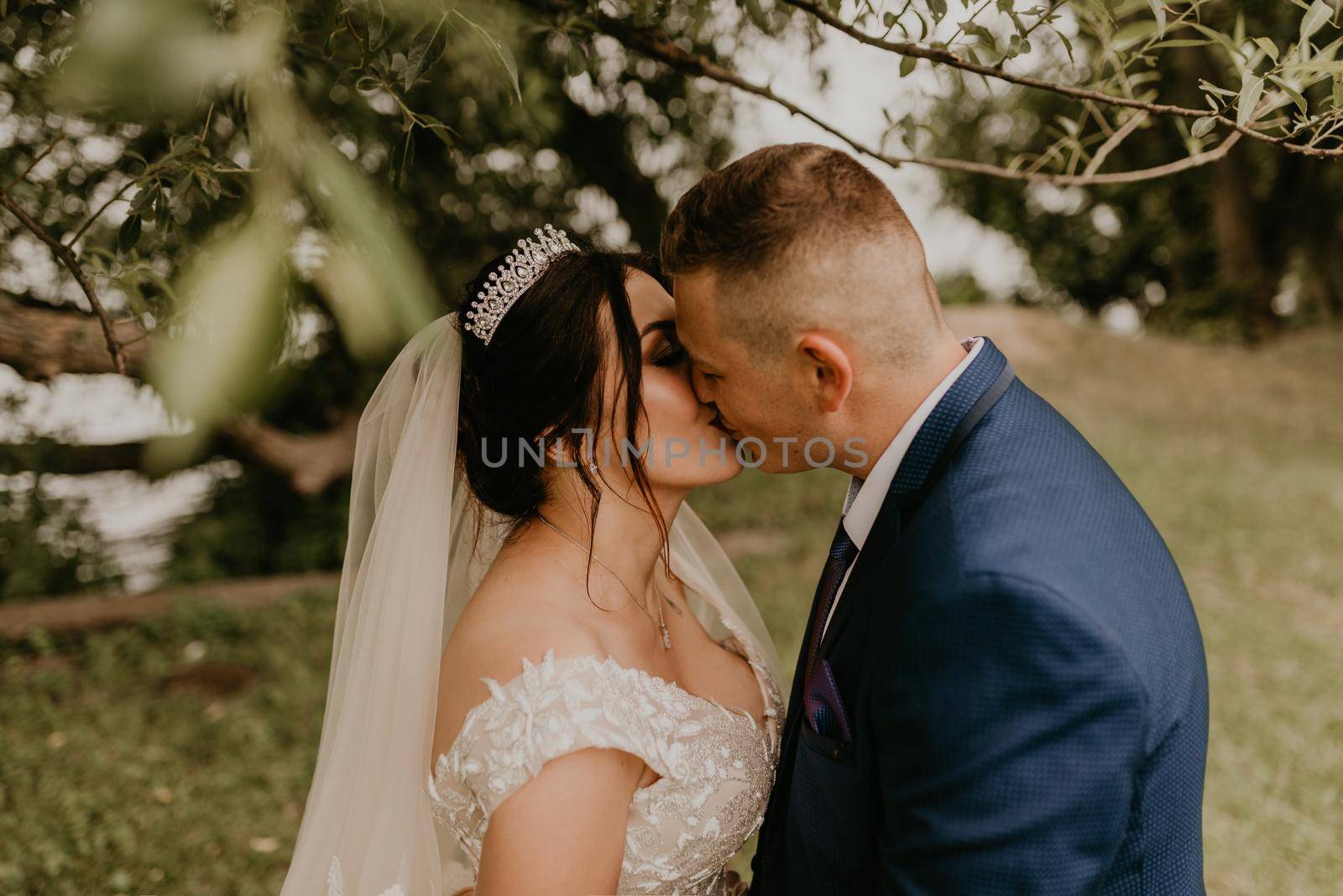 blonde European Caucasian young man groom in blue suit and black-haired woman bride in white wedding dress with long veil and tiara on head. Newlyweds kiss under a tree in summer